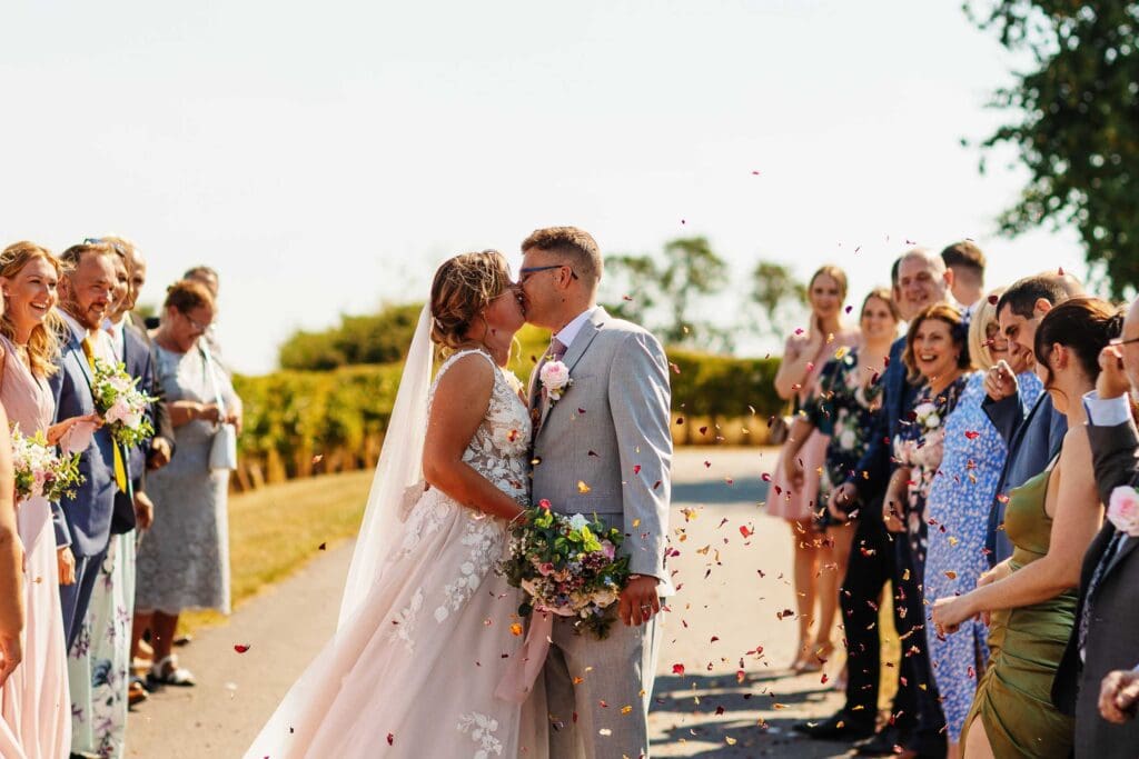 Elegant couple sharing a kiss with wedding guests throwing confetti on a sunny outdoor wedding day, captured at BGS Weddings, showcasing romantic wedding celebrations and joyful moments.