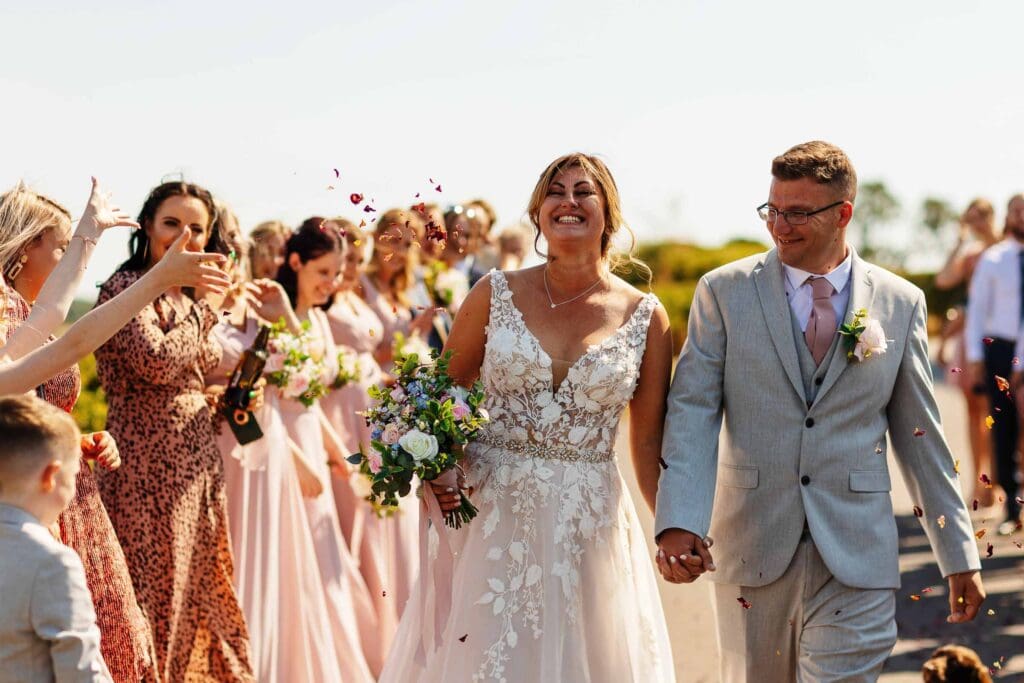 Vibrant outdoor wedding celebration with a happy bride and groom walking hand in hand as guests throw colourful confetti during the ceremony.