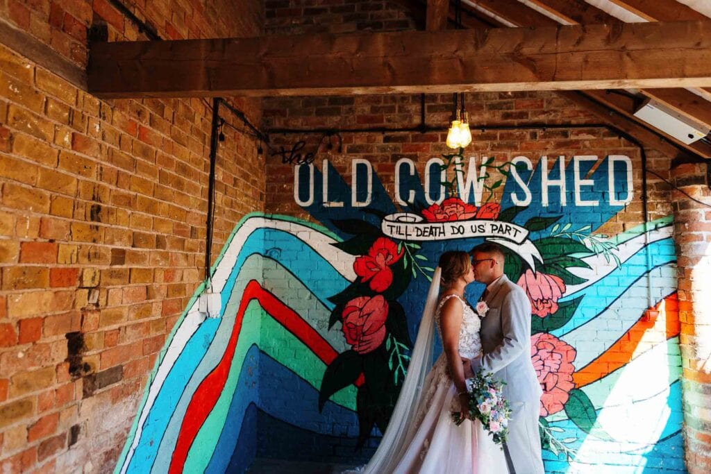 Colourful brick-walled wedding venue with a mural backdrop, featuring a bride and groom sharing a kiss at The Old Crown Shed, perfect for rustic weddings and unique wedding photography.