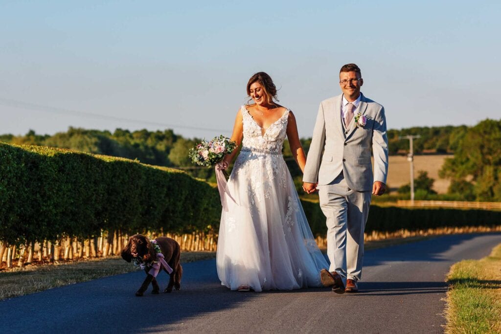 Elegant bride and groom walking hand in hand on a scenic countryside wedding day, with a Labrador dog in a bowtie, celebrating love and wedding vows at BGS Weddings, UK.