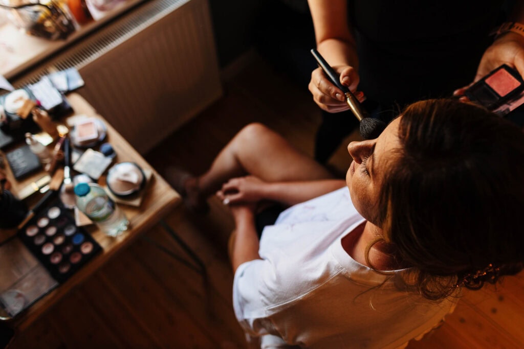 Makeup artist applying foundation to bride before her wedding, with makeup products and tools on a wooden table.