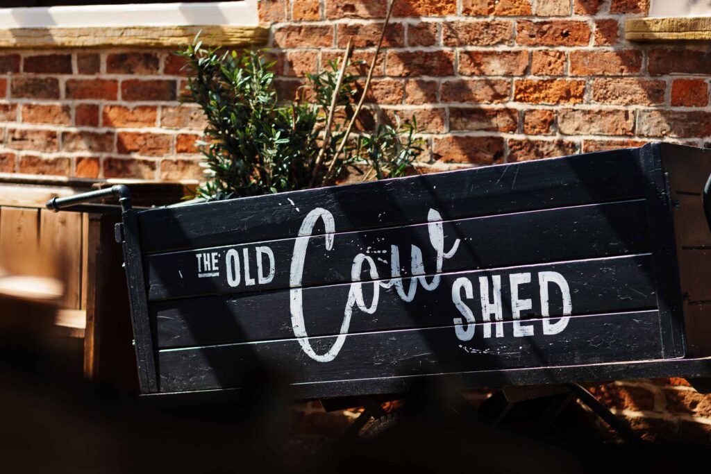 Vintage wooden garden shed with "The Old Coxy Shed" painted on it in white letters, set against a brick wall with potted plants. Perfect for rustic wedding themes and outdoor events.