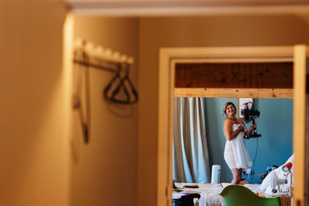 Elegant bride preparing for wedding, smiling while holding a fan, captured in a mirror reflection at BGS Weddings. Wedding photography, bridal makeup, and hairdressing services offered in the UK.