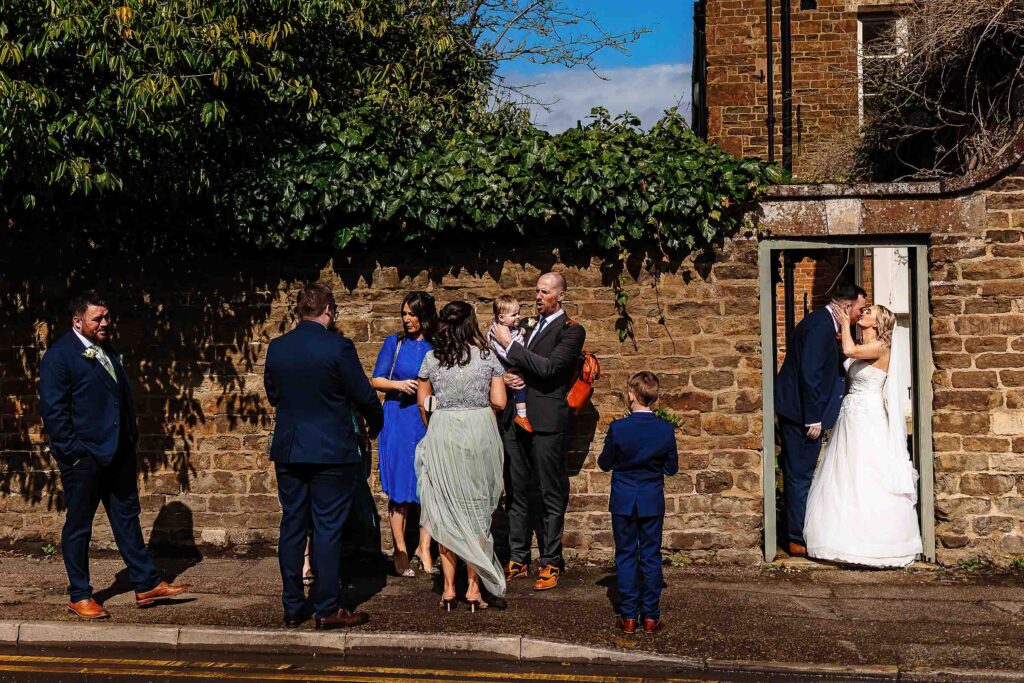 Elegant wedding couple sharing a kiss in the doorway of a rustic brick building, with wedding guests engaging outdoors, capturing lovely wedding moments and scenic backgrounds.
