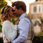 Romantic couple sharing an intimate moment outdoors during a wedding, with a historic building in the background, captured in warm golden hour light.