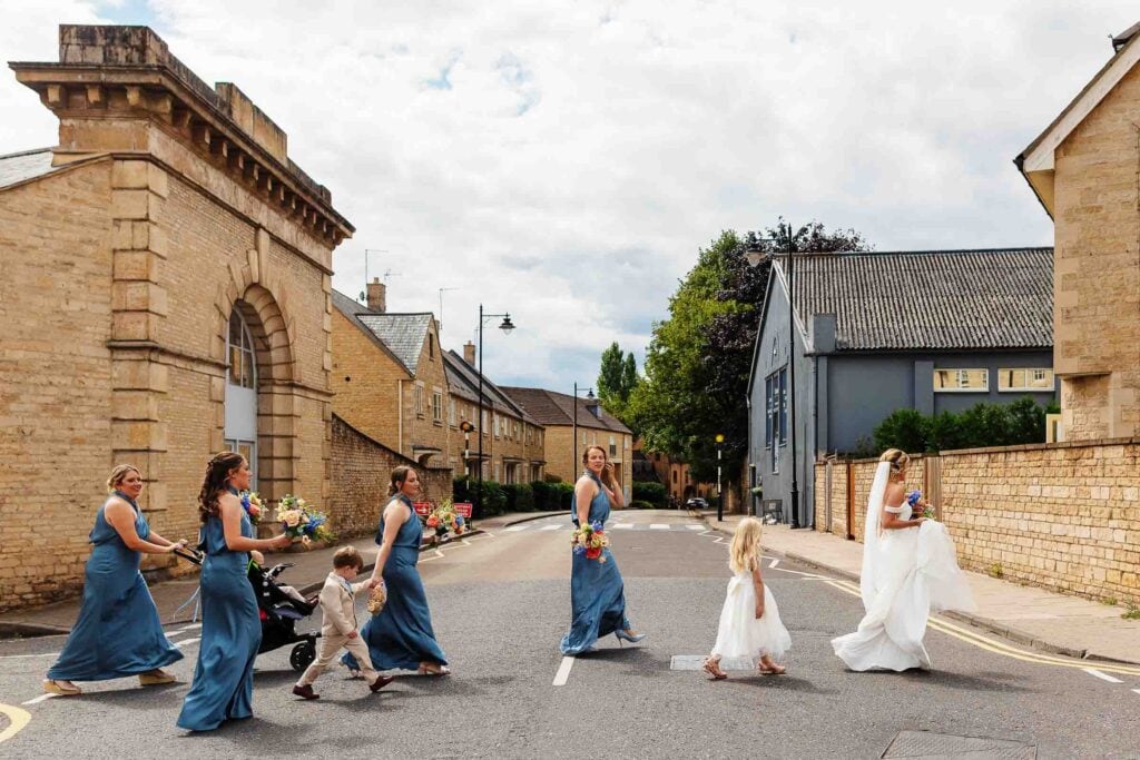 Elegant wedding procession crossing a quaint UK village street, bridal party in blue dresses, bride in white, groom and children participating, charming historic architecture, perfect for wedding photography.