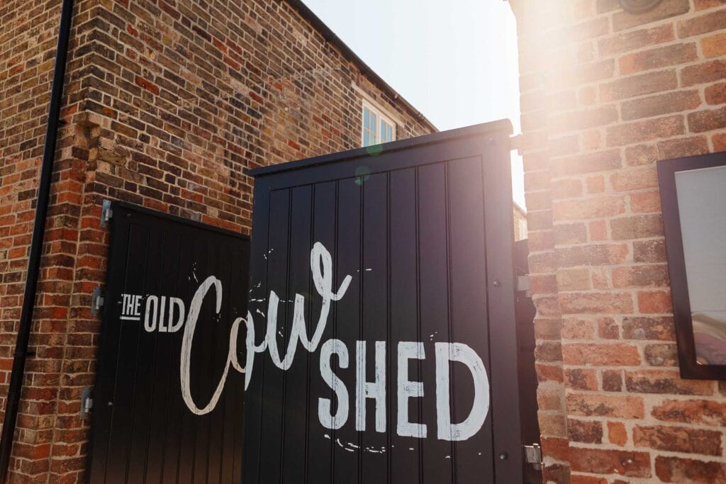 Rustic brick exterior of the Old Cow shed wedding photographer venue with a black wooden door displaying the venue's name, bathed in warm sunlight for an inviting wedding setting.