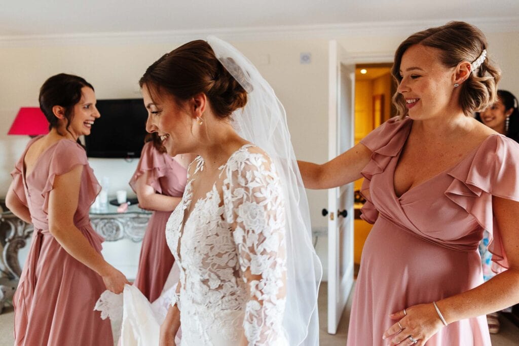 Elegant bride in intricate lace wedding gown getting ready with her bridesmaids in dusty pink dresses at BGS Weddings.
