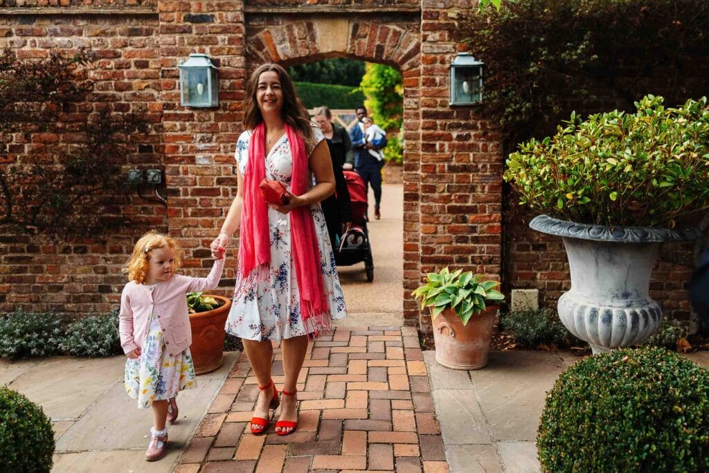 Enjoying a sunny day at a wedding venue, a smiling woman in a floral dress and red scarf walks hand in hand with a young girl near a brick archway surrounded by potted plants, perfect for wedding celebrations.