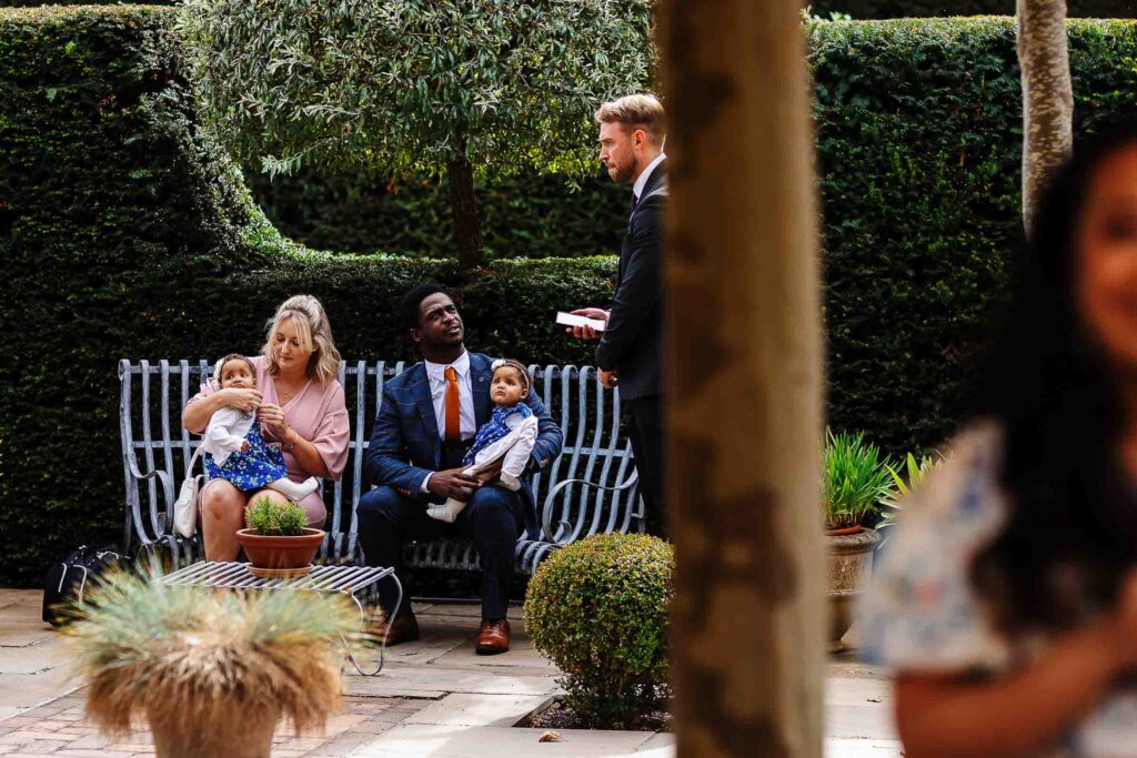 A professional outdoor wedding ceremony with a diverse group of guests, including children, seated on a bench in a lush garden setting, with a officiant conducting the vows.