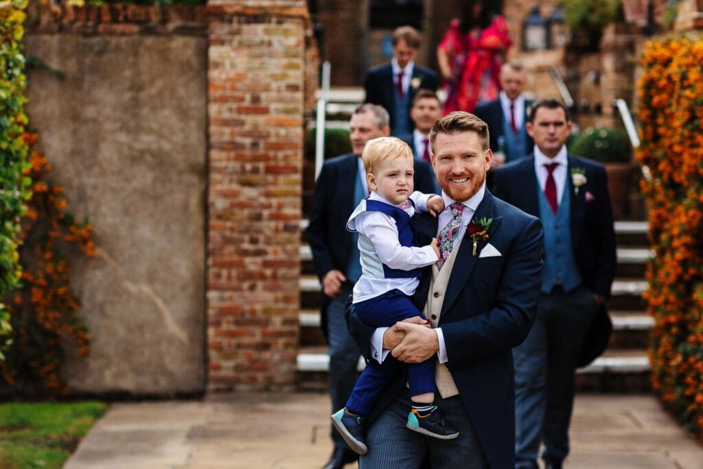 Elegant groom holding a young ring bearer during a wedding ceremony outside, with groomsmen in suits and colorful flowers, set against historic brick and stone architecture.