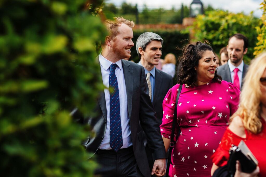 Elegant wedding guests walking outdoors, dressed in formal attire, enjoying the beautiful garden setting for a wedding celebration at BGS Weddings.