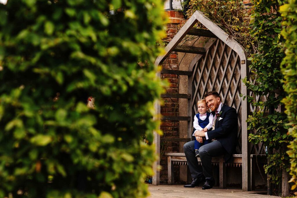 Elegant wedding photography featuring a groom and a young boy sitting on a wooden garden bench under an arbor, surrounded by lush greenery, perfect for wedding venues and outdoor wedding events.