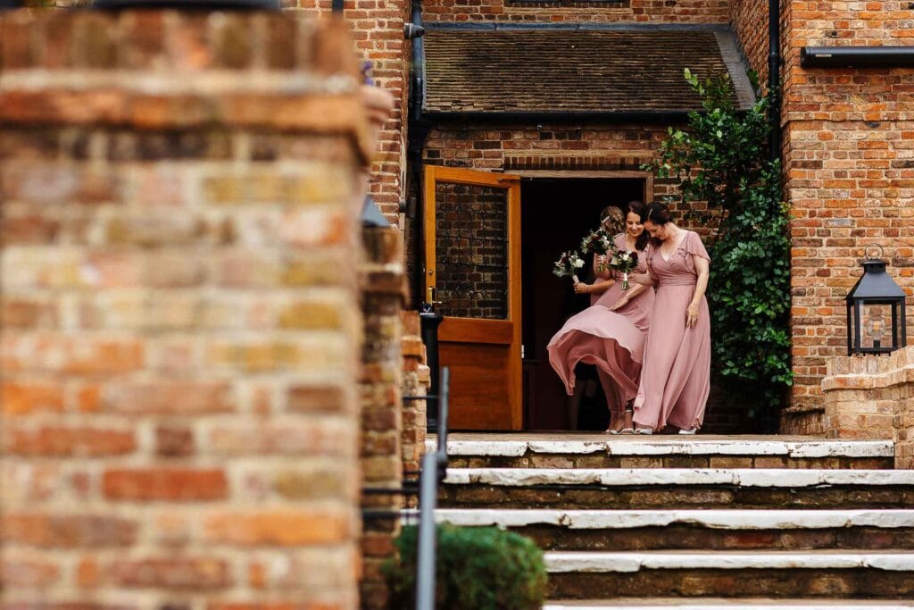 Elegant bridesmaids in blush pink dresses with floral bouquets exiting a rustic brick wedding venue.