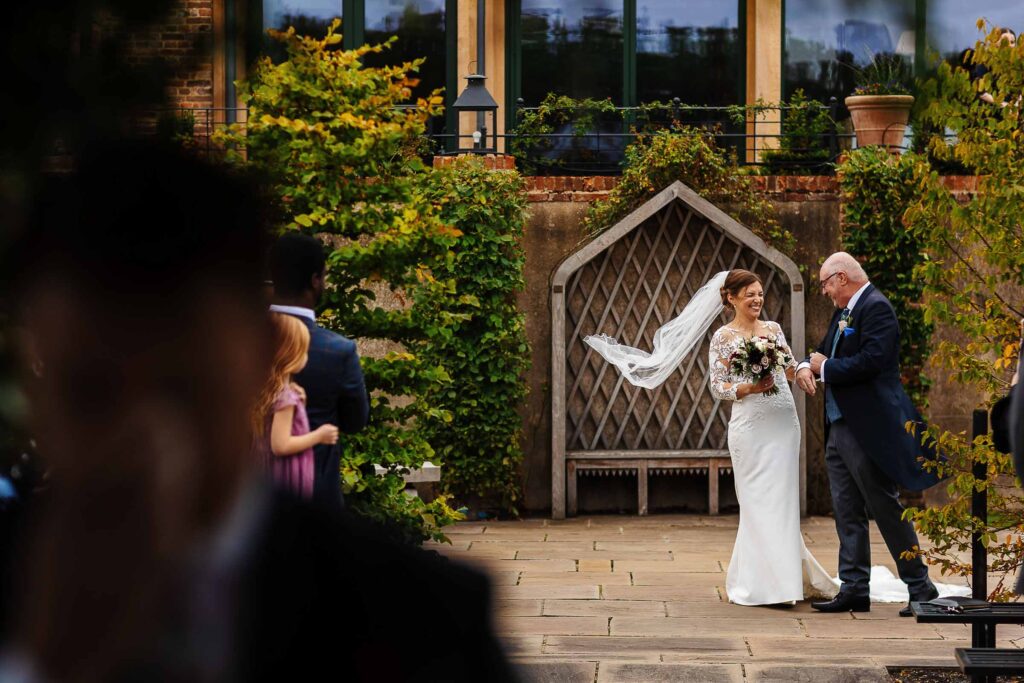 Elegant bride in a lace wedding dress with a bouquet, smiling as she exchanges vows with her father during an outdoor wedding ceremony at BGS Weddings, surrounded by lush greenery.