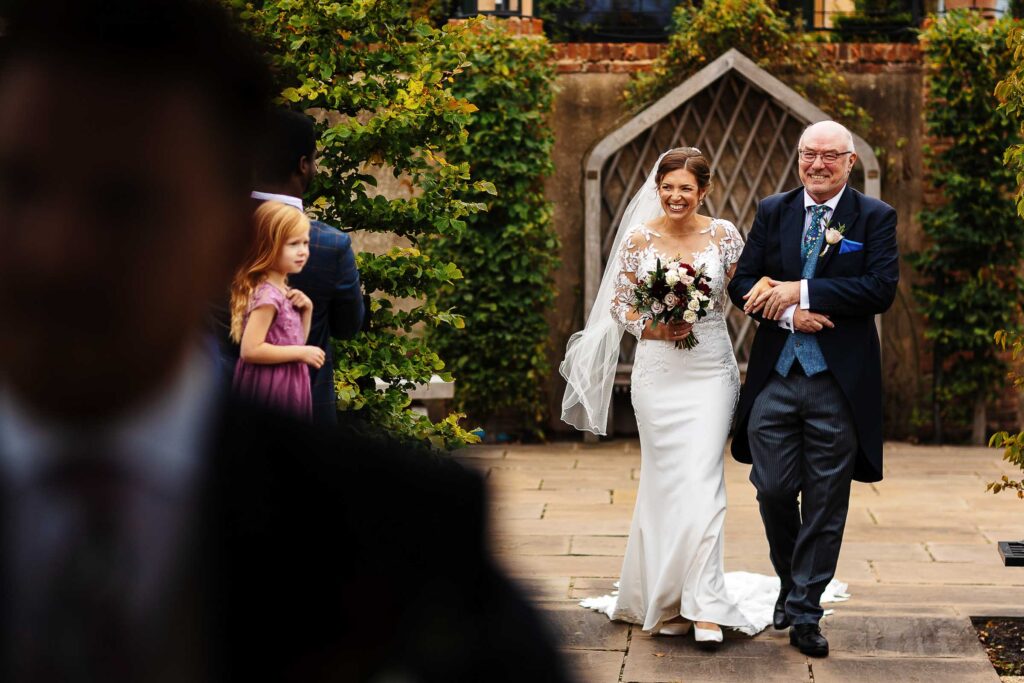 Elegant bride walking down the garden aisle with her father during a wedding ceremony, surrounded by lush greenery, capturing a joyful moment for BGS Weddings.