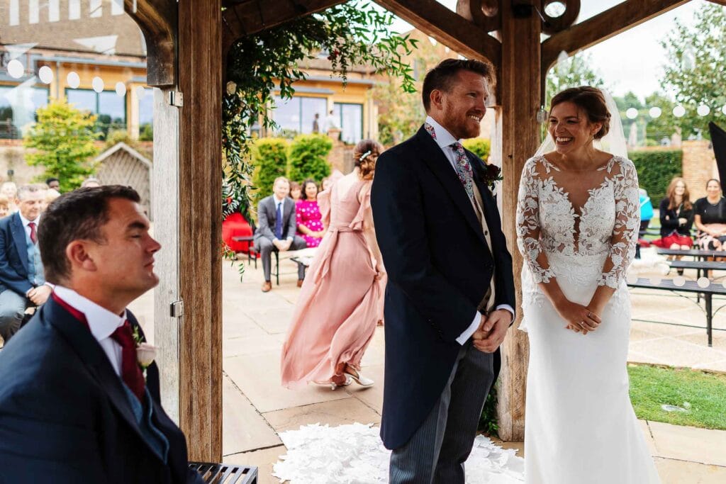 Elegant outdoor wedding ceremony at BGS Weddings in the UK, featuring a smiling bride and groom exchanging vows under a rustic wooden gazebo with guests in the background.