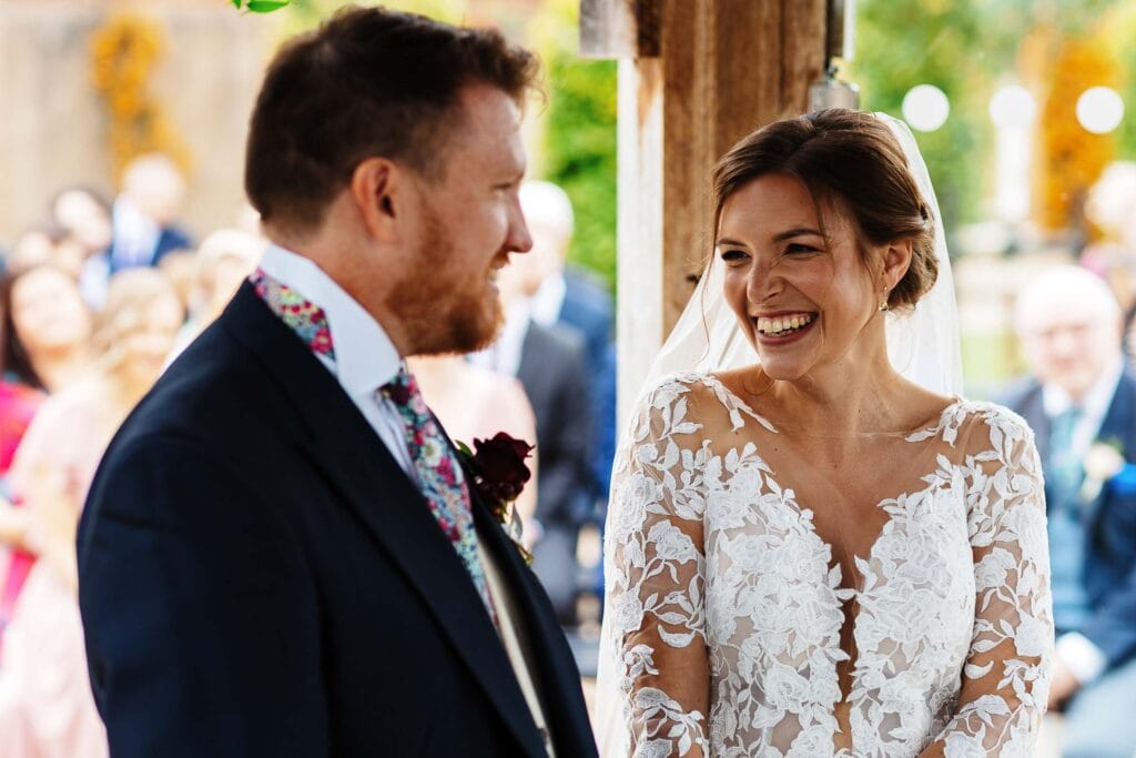 Romantic outdoor wedding ceremony with bride and groom smiling, featuring bride in lace wedding dress and groom in a floral tuxedo, surrounded by guests at BGS Weddings UK.