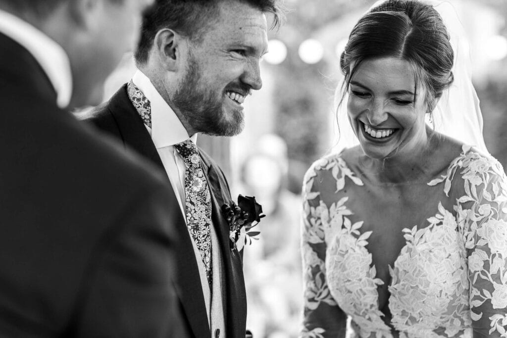 Joyful bride and groom during wedding ceremony, exchanging vows and sharing a happy moment, captured in black and white, showcasing love and celebration at BGS Weddings.