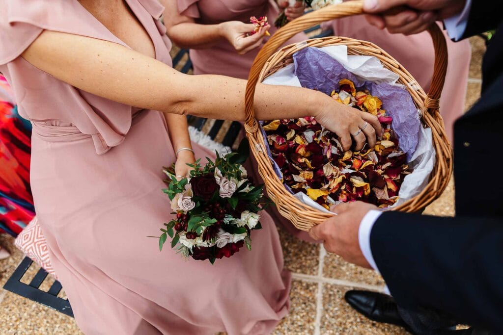 Rose petal confetti being handed out to guests at a wedding ceremony, with bridesmaids in pink dresses and a bride holding a bouquet, capturing a joyful and romantic moment.