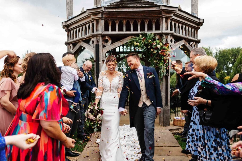Elegant wedding ceremony at BGS Weddings showcasing a joyful bride and groom walking under a floral decorated wooden arch, surrounded by friends and family celebrating their special day.