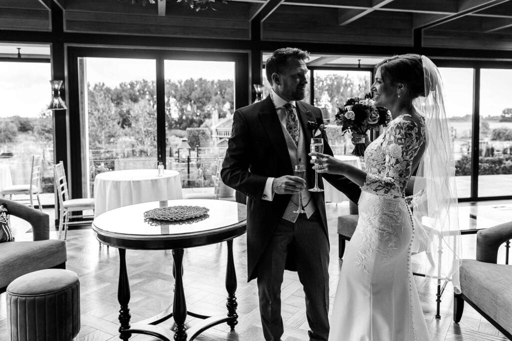 Elegant bride and groom celebrating their wedding joyfully indoors at BGS Weddings venue, holding champagne glasses, bright natural light, romantic atmosphere, stylish wedding dress, classic suits, modern decor, scenic view outside, celebration, happiness.