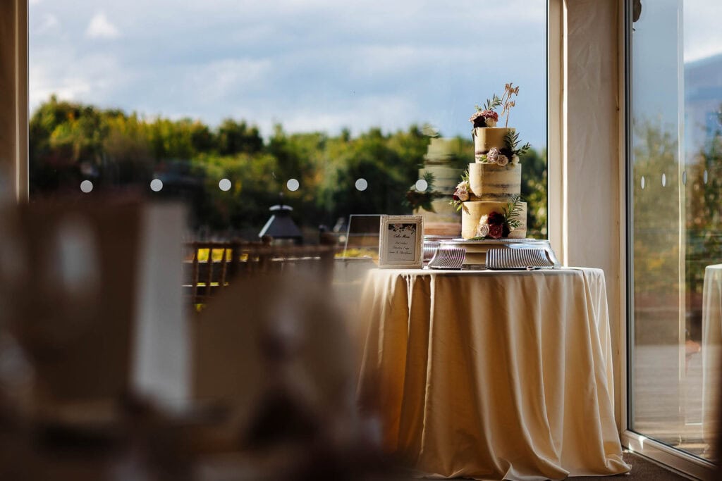 Elegant wedding cake displayed on a round table with cream tablecloth at BGS Weddings venue, featuring natural light and scenic outdoor views for a perfect wedding celebration.