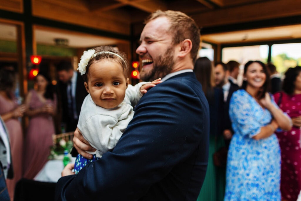 Smiling man holding a young girl at a wedding reception, joyful atmosphere, wedding celebration, BGS Weddings UK.