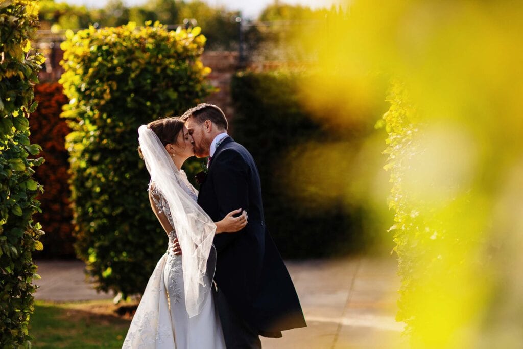 Elegant couple sharing a romantic kiss outdoors during a wedding photography session, surrounded by lush greenery and vibrant autumn foliage.