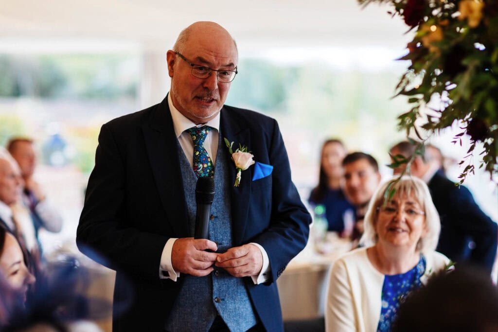 Elegant man giving speech at wedding reception, holding a microphone, dressed in a navy suit with a floral tie, surrounded by family and friends celebrating the wedding day.