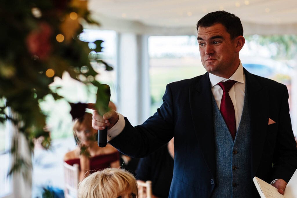 Elegant groom giving a speech at wedding reception, dressed in a tailored navy suit with a red tie, holding notes and microphone, with guests and floral decorations in the background.