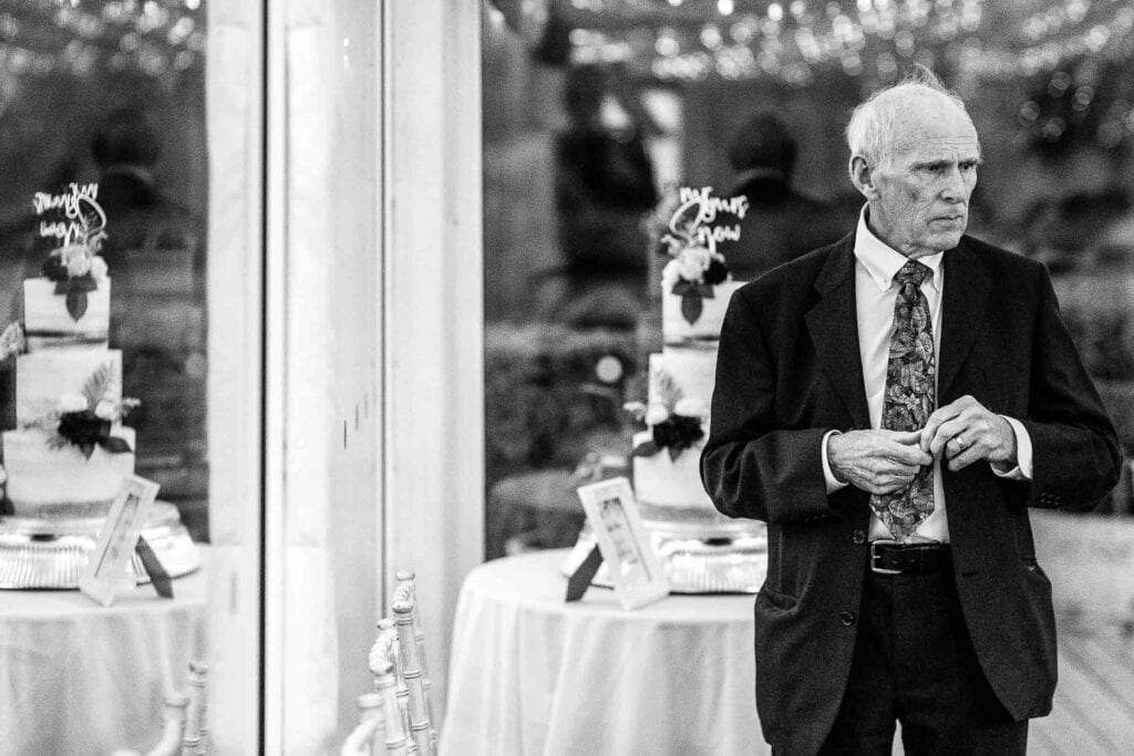 Elegant elderly man in a suit at a wedding reception, standing near decorated tables with wedding cakes, capturing a moment of reflection and solemnity during the celebration.