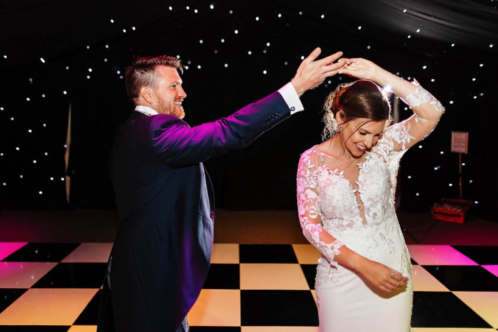 Elegant bride and groom sharing a joyful moment on the dance floor at a wedding reception, showcasing celebration, love, and happiness with a stylish black and white checkered dance floor and stunning backdrop.