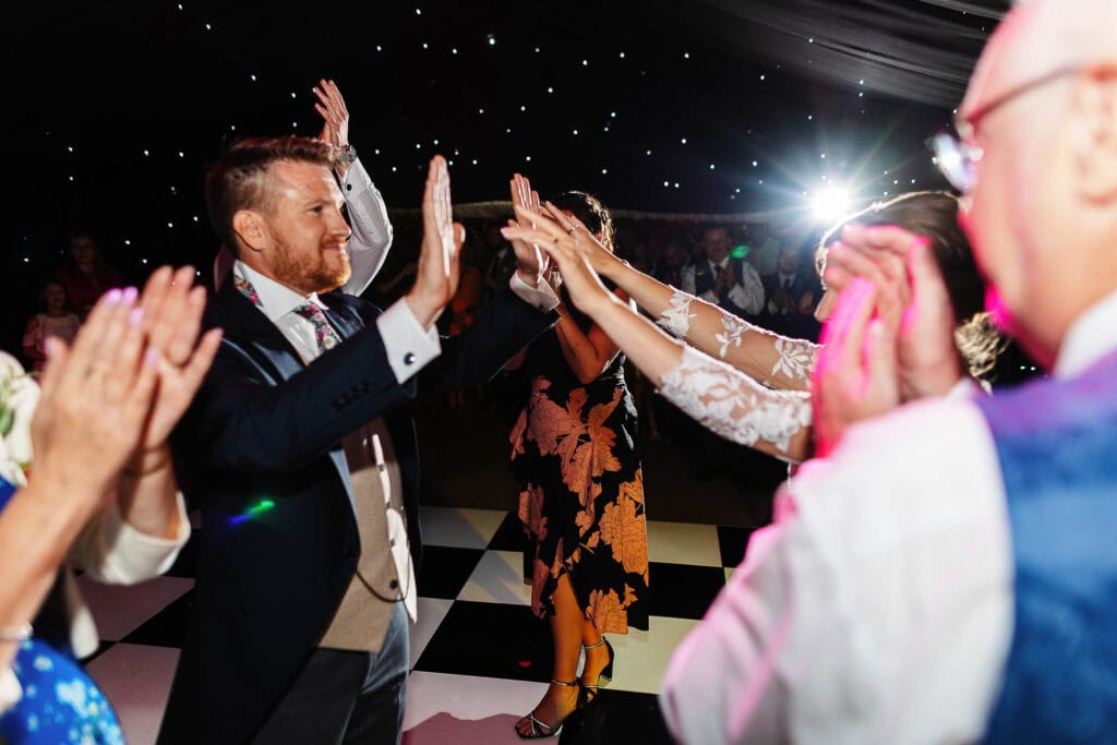 High-energy wedding celebration with guests dancing and high-fiving at BGS Weddings in a stylish venue with a starry ceiling and black-and-white checkered dance floor.
