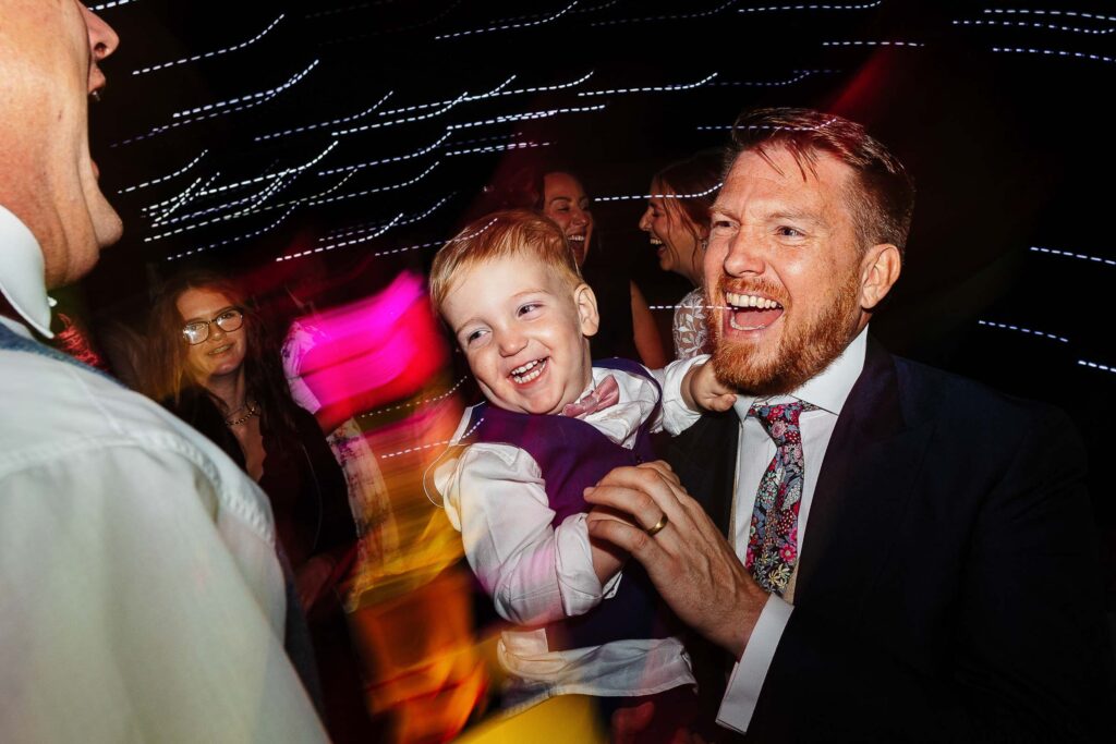 Joyful wedding celebration with smiling guests and dancing under colourful party lights at BGS Weddings.
