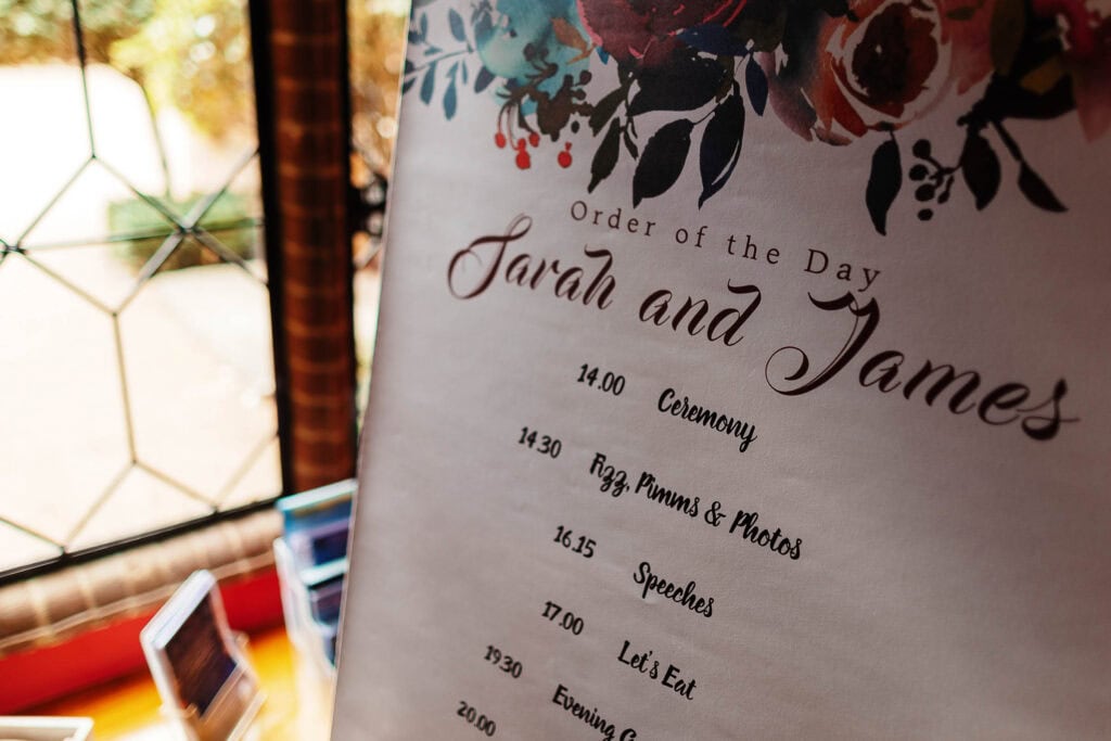 Elegant wedding schedule sign for Sarah and James, featuring time slots for ceremony, speeches, meal, and evening entertainment at a picturesque outdoor venue with floral decor.