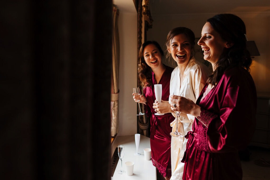 Elegant bridal party preparing in satin robes, celebrating before the wedding, holding champagne glasses, in a sophisticated, warmly-lit room at BGS Weddings.