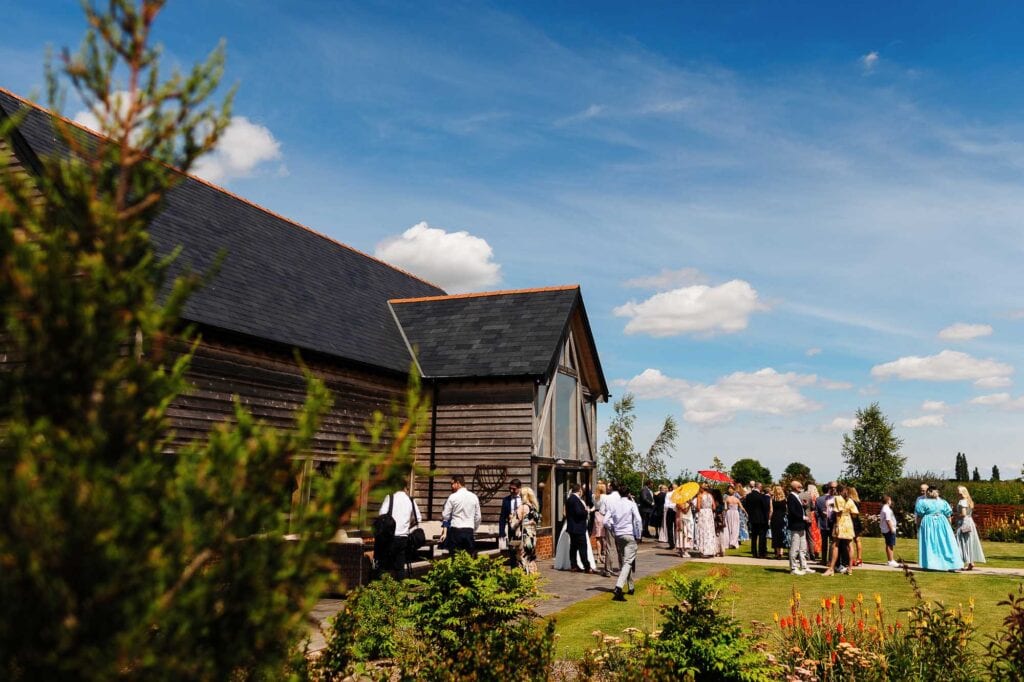 People gathering for outdoor wedding at BGS Weddings venue with rustic barn, lush gardens, and sunny sky, creating a perfect setting for couples' special day.