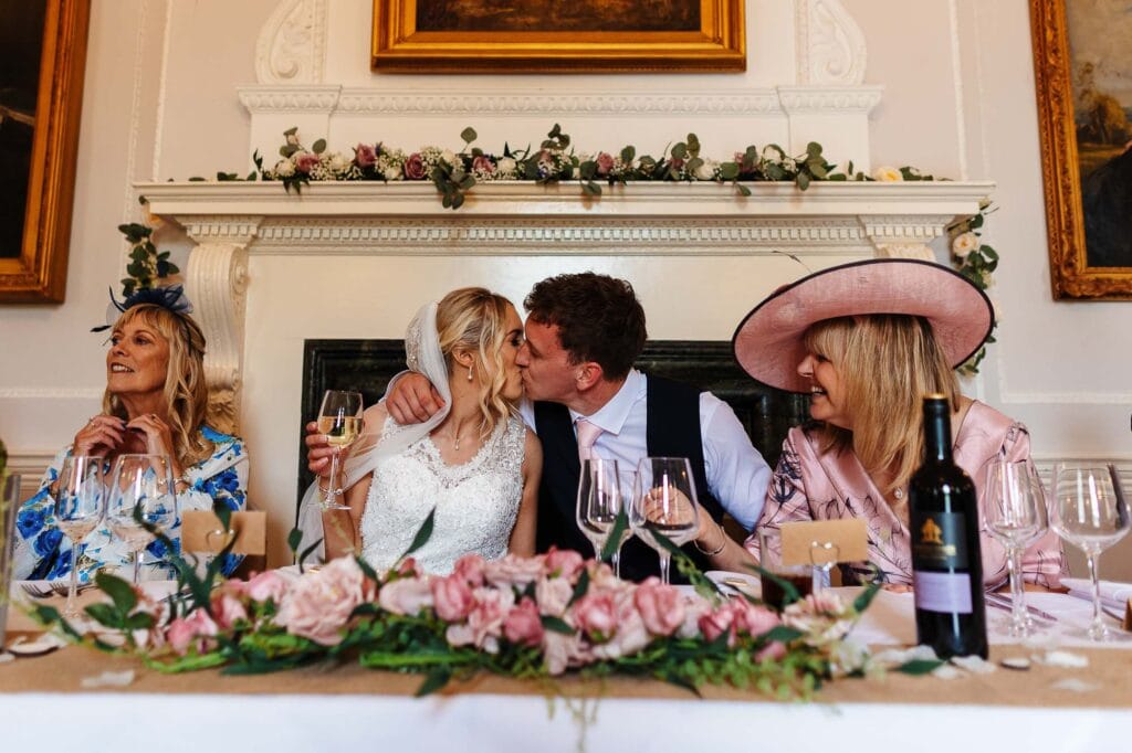 Elegant wedding reception with newlywed couple sharing a kiss at a beautifully decorated table in a historic venue. Guests are enjoying the celebration with floral arrangements and fine wine.