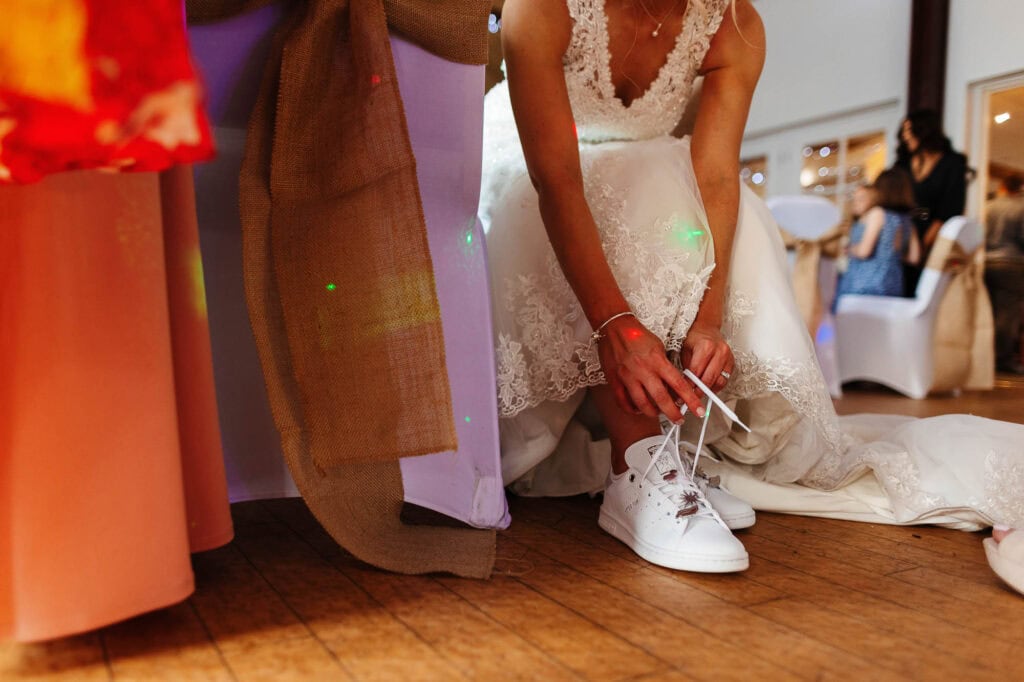 Elegant bride in lace wedding dress tying white wedding shoes at reception, capturing special wedding day moments with wedding cake and guests in the background.
