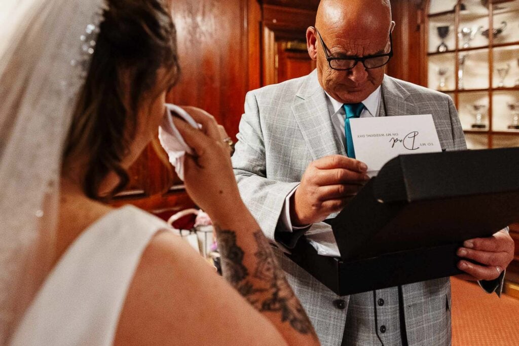 Brooksby hall wedding photographer man in a light grey suit presenting a wedding gift to bride with tattoos in a boutique wood interior.