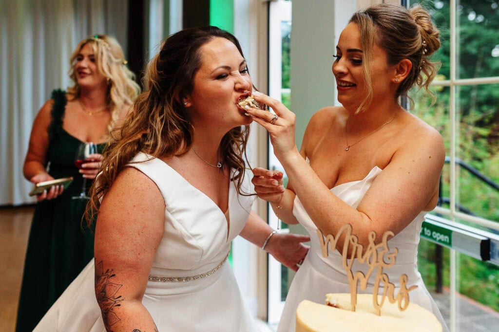 Elegant bride enjoying cake at wedding reception, surrounded by joyful friends, with natural light and green outdoor views in the background.