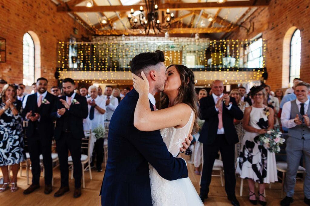 Elegant wedding couple sharing a kiss in a rustic barn wedding venue with exposed brick walls, fairy lights, and a joyful audience celebrating in the background.