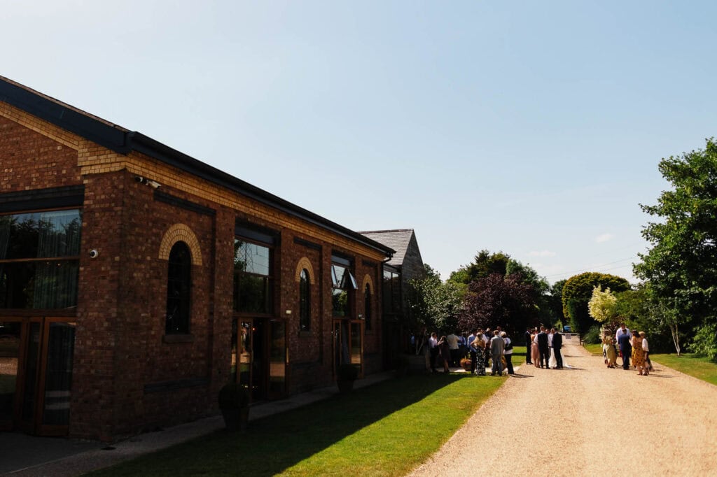 Elegant brick wedding venue with outdoor garden setting, outdoor gathering for wedding reception, lush green trees and clear sky, perfect for weddings at BGS Weddings UK.