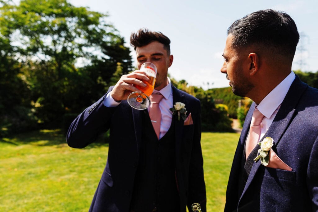 Gentlemen in navy suits celebrating at an outdoor wedding with pink accents, drinking and socialising on a sunny day, with greenery in the background.