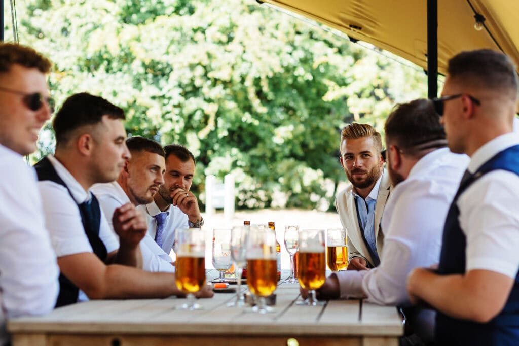 Relaxed outdoor wedding reception with groomsmen enjoying drinks and conversation under a canopy, surrounded by lush greenery. Perfect setting for a stylish and memorable wedding celebration.