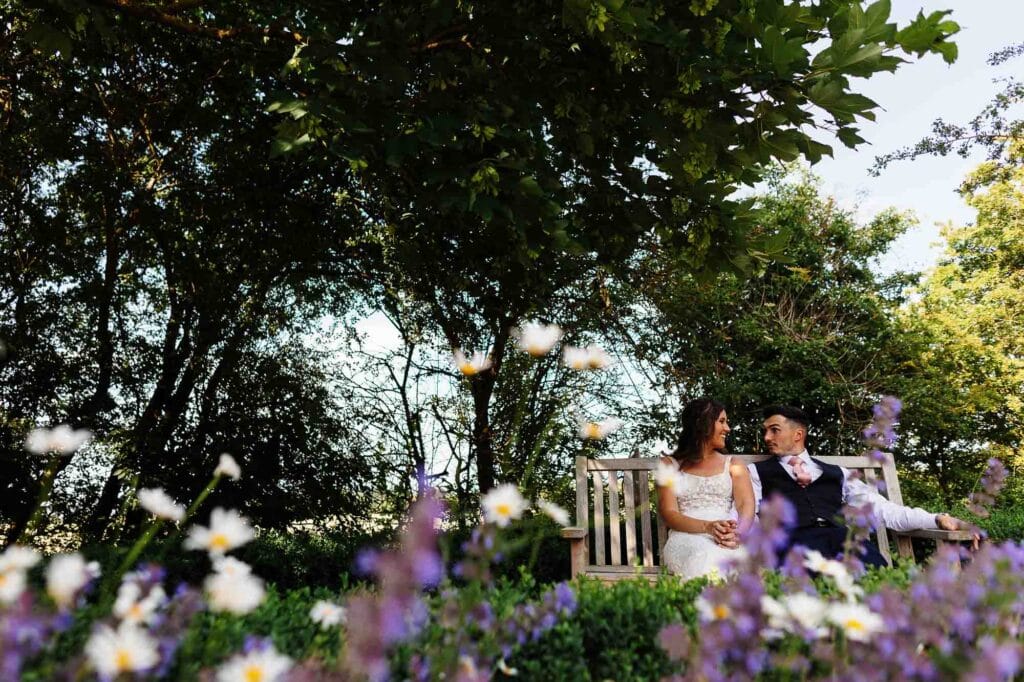 Romantic wedding couple sitting on a wooden bench amidst lush greenery and vibrant flowers, capturing a beautiful outdoor wedding scene for BGS Weddings.