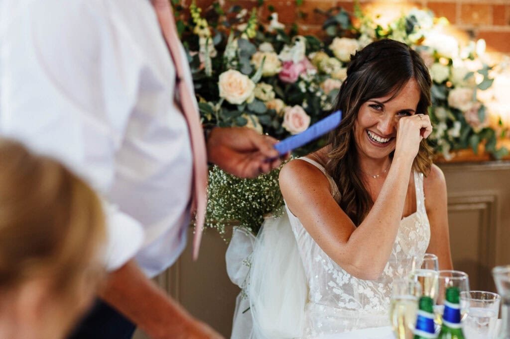 Elegant bride laughing during her wedding speech, surrounded by floral decor at a BGS Weddings event in the UK.