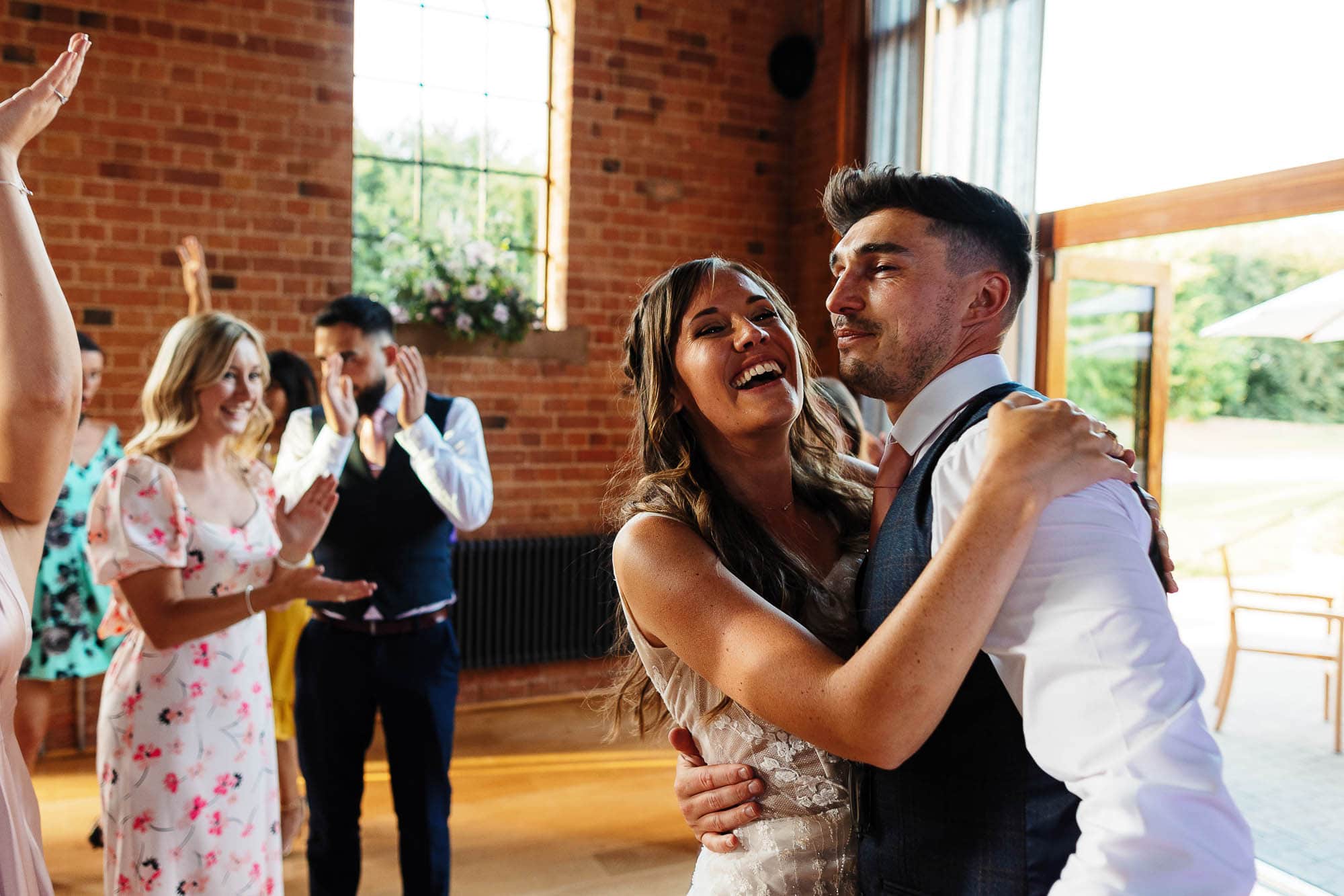 Elegant couple dancing at wedding reception in rustic brick venue, surrounded by joyful guests, celebrating love and happiness, captured by BGS Weddings.