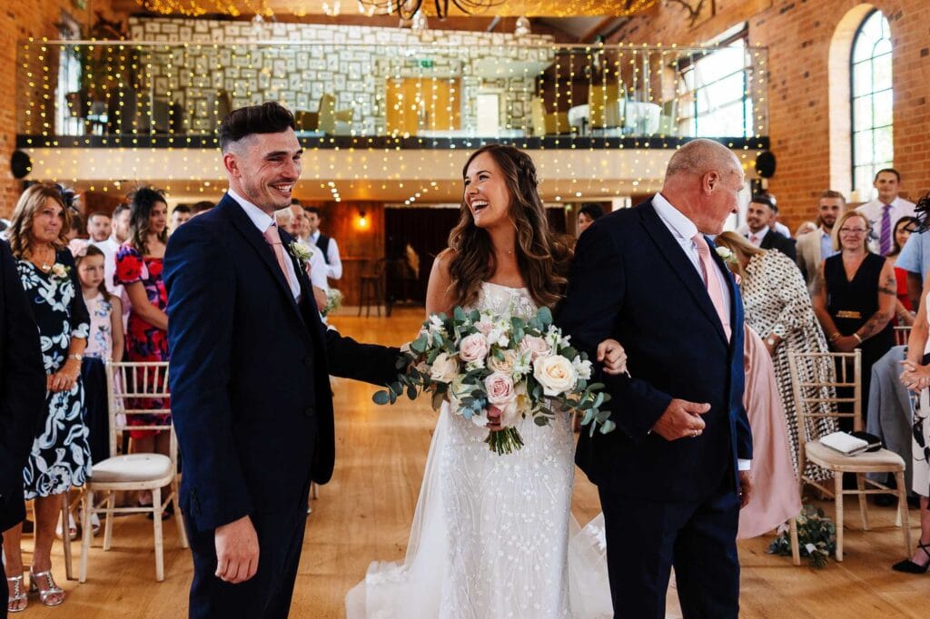 Elegant bride walking down the aisle with her father, surrounded by happy wedding guests in a stylish rustic venue featuring warm lighting and floral decor.